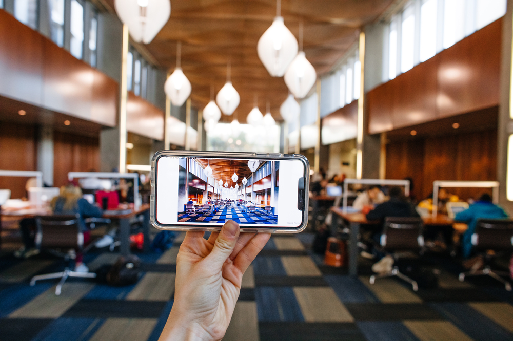 In the UCSB Library, a hand holds up a smartphone displaying a photo of the room.